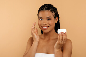 Beautiful Young Black Female Applying Moisturising Cream On Face While Standing Against Beige Background, Attractive African American Woman Holding Jar With Nourishing Lotion And Smiling At Camera