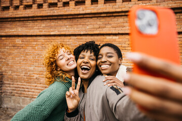 Three cheerful female tourists are taking a selfie with a smartphone in front of a brick wall,...