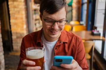 Happy man enjoying a break in a pub, sipping a cold beer while using his smartphone. Smiling and relaxed, he browses social media on his mobile device, embracing leisure time