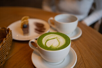 Warm matcha latte with a heart design resting on a wooden table in a cafe, accompanied by a small dessert and another beverage, creating a cozy atmosphere