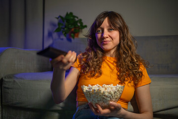 Happy young woman enjoying popcorn while watching television, sitting comfortably on a cozy sofa at...