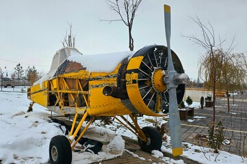Abandoned yellow propeller plane in a snowy park. The aircraft features a single propeller at the front and visible damage exposing its framework. The yellow paint is faded, with snow covering section