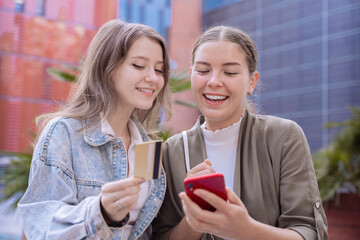 Two smiling Caucasian women using a smartphone and credit card for online shopping outdoors. Modern lifestyle, digital payment, and technology concept in an urban setting with natural daylight.