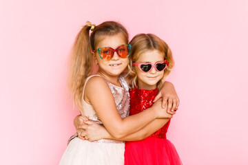 Two cheerful girls in sunglasses hugging on a pink background