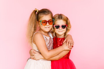 Two cheerful girls in sunglasses hugging on a pink background