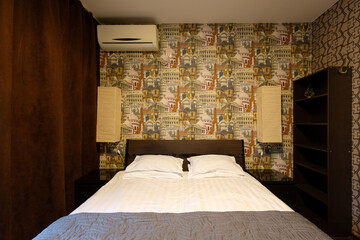 Apartment interior. Bedroom. Double bed in the bedroom. Pillows and bedspread on the made bed. Wallpaper with images of European landmarks. Air conditioning and wall lamps on the wall.