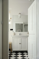 Vintage elegant bathroom interior through open door. Wash basin, cabinet, mirror, glass pendant. Black white checkered floor, white tile wall background. Elegant minimal Scandinavian style design