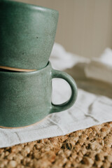 Two green ceramic coffee cups are sitting on a white cloth. The cups are placed on a brown rug, which adds a warm and cozy touch to the scene. Scene is inviting and comfortable