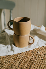 Two cups are sitting on a table with a white cloth underneath them. The cups are brown and have a rustic, earthy feel to them. The scene is simple and peaceful, with the cups