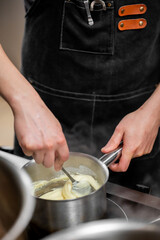 A chef stirs a creamy mixture in a saucepan on the stovetop. They wear a black apron with leather...