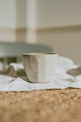 A white ceramic cup with a brown rim sits on a table. The cup is empty and has a simple design. The table is covered with a white cloth, and there is a chair in the background. The scene is calm