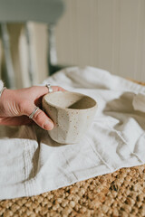 A hand holding a white ceramic cup on a white cloth. The cup is small and has a unique design. The scene is simple and peaceful, with the focus on the cup and the hand holding it