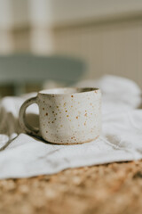 A white ceramic coffee cup with a brown speckled design sits on a white cloth. The cup is placed on a wooden table, and the cloth is spread out on the table. Concept of warmth and comfort