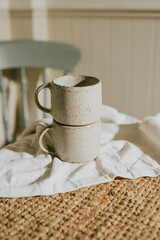 Two white ceramic coffee cups sit on a tablecloth. The cups are placed on top of a woven rug. Concept of warmth and comfort, as the cups are likely to be used for enjoying a hot beverage
