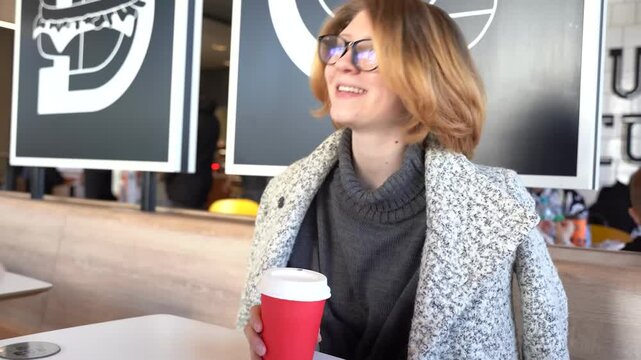Young smiling happy young woman in glasses sit at table in cafe shop, drink coffee and speak another