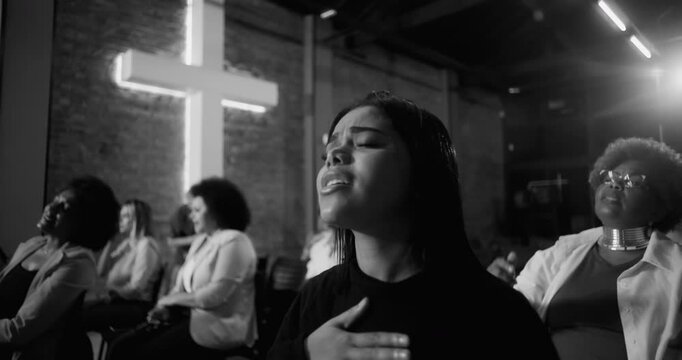 Young woman singing in prayer during worship service, hand on chest in devotion, illuminated cross and congregation in background, black and white tones