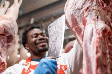 Smiling butcher working in slaughterhouse cutting hanging raw meat.