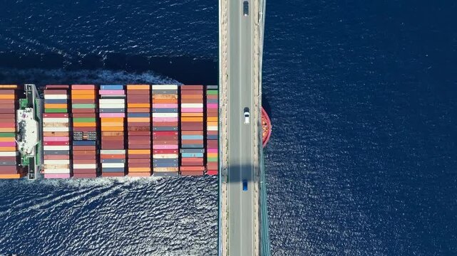 Transport concept with an aerial overhead view of a very big container super cargo ship passing under a bridge with car traffic