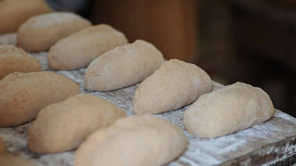Close-up of rows of unbaked dough rolls resting on a floured wooden board, showing their texture and shape before baking, evoking traditional bread-making methods in a rustic setting.