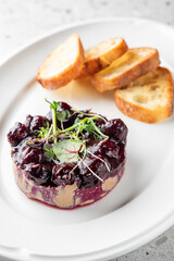 A gourmet dish featuring a rich, dark berry compote served over smooth paté, garnished with microgreens, accompanied by slices of toasted bread on a white plate.