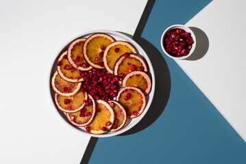 Rodajas de naranja sanguina y semillas de granada en un plato blanco sobre un fondo blanco y azul....
