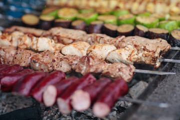 Close-up view of various skewers grilling over charcoal, featuring seasoned meats, sausages, and a mix of green zucchini, eggplants, and yellow peppers, creating vibrant assortment of grilled flavors