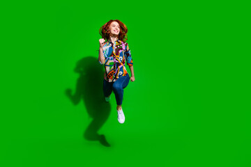 Joyful young woman jumping with laughter against a bright green backdrop wearing trendy vibrant printed shirt and casual jeans