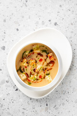 Creamy soup with croutons, garnished with green and red sauces, served in a white bowl on a white plate with a napkin, placed on a stone surface. Vibrant and appetizing presentation.