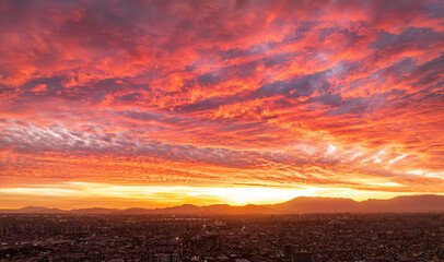 Santiago, Chile, at sunset, with a vibrant orange, pink, and red sky streaked with clouds, illuminating the cityscape as lights begin to glow in the transition from day to night.