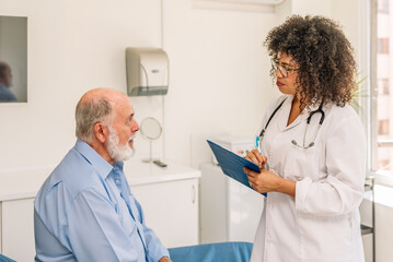 Female Afro doctor taking notes while listening to senior patient in hospital room