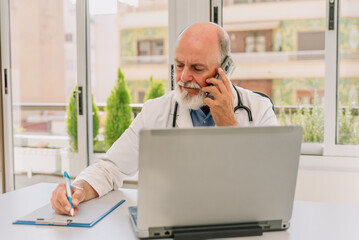 Senior doctor in lab coat sitting at desk talking on the phone and taking notes in medical office