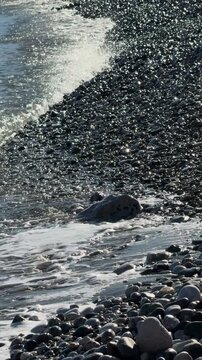 CLOSE UP, VERTICAL: Powerful wave crashes onto pebbled shore with sea foam and water droplets flying. Sunlight glistens on smooth stones, creating sparkling texture. Coastal energy and natural beauty