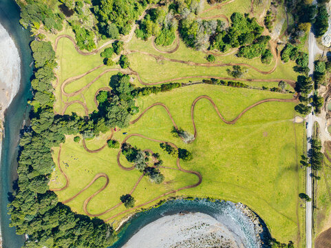 Aerial view of a motorcycle circuit in Puerto Fonk, Los Lagos, Chile, with winding dirt tracks on a grassy field, surrounded by trees and a river under a clear sky.