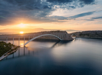 Naklejka premium Aerial view of bridge and blue sea at colorful sunset. Modern Krk bridge island during golden hour, mountains, rocks, reflection in water and cloudy sky. Top drone view of bridges. Architecture