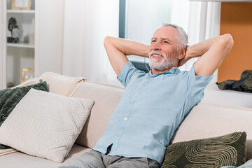 Fototapeta premium Smiling retired caucasian senior mature middle aged man relaxing on sofa in the living room at home, enjoying sunny afternoon. Lifestyle happy retirement concept