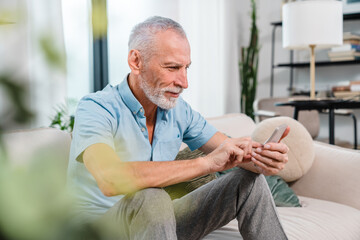 Happy smiling senior man using smartphone device while sitting on sofa at home. Mature man lying on couch reading messages on mobile phone, relaxing at home.