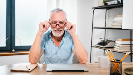 Smiling older mature middle aged man businessman wearing eyeglasses looking at camera sitting at table, using laptop working online remotely, e-learning, browsing web, searching online at home office