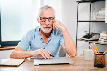 Close up portrait of Smiling older mature middle aged professional man freelancer looking at camera sitting at table, using laptop working, browsing online relaxing at casual home office .