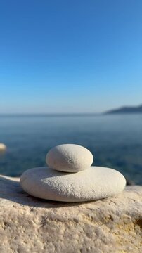 CLOSE UP, VERTICAL: Flying rock hits and collapses a balanced white stone tower by the sea. Smooth pebbles start to fall against calm ocean backdrop and cloudless sky. Summer shenanigans at the beach.
