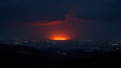 A nighttime panoramic view showing a city's lights against a dark sky with clouds and a wildfire burning brightly on the horizon.