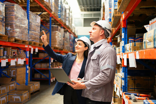 Two professionals inspect inventory in a warehouse, discussing operations and logistics while using a laptop for data analysis.