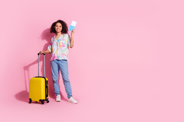Young woman with curly hair holding a passport and standing with a suitcase against a pink...