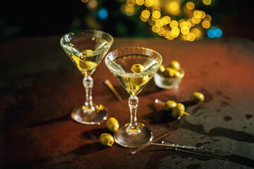Two martini glasses filled with clear liquid and green olives stand on a table with scattered olives, while golden bokeh lights in the background create a warm holiday ambiance.
