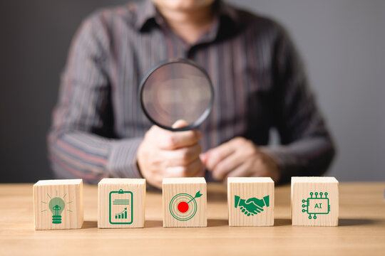 businessman examines business strategy concepts with a magnifying glass, focus on wooden blocks display icons for innovation, analytics, goal setting, partnerships, and AI.