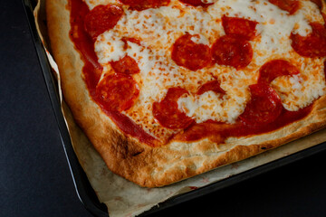 A square pepperoni pizza with ham and pineapple, presented on a black background in a dark food style. The soft light highlights the texture of the artisanal crust, melted cheese, and ingredients