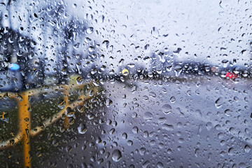 Raindrops on a glass window with a blurred urban street view. Wet surface on a rainy day texture. Moody city atmosphere seen through a bus stop window in cloudy weather.