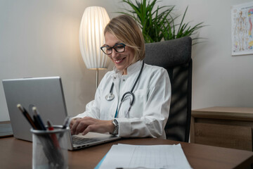 Mature Caucasian female doctor in white medical uniform sit at desk in hospital work on laptop make note in register. Woman nurse or GP fill patient anamnesis in journal, use computer
