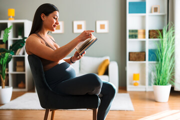 Pregnant woman relaxing on chair reading a book in her modern living room, enjoying quiet time