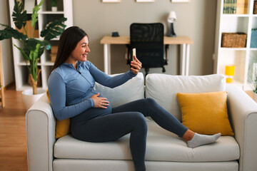 Obraz premium Happy pregnant woman making video call with smartphone while sitting on comfortable sofa at home