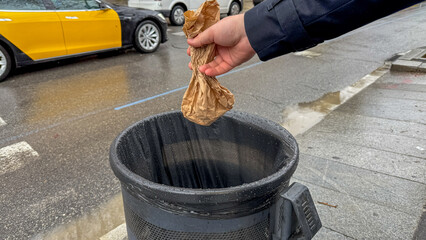 A male hand discards a crumpled paper bag into a trash can on a rainy street, promoting urban...